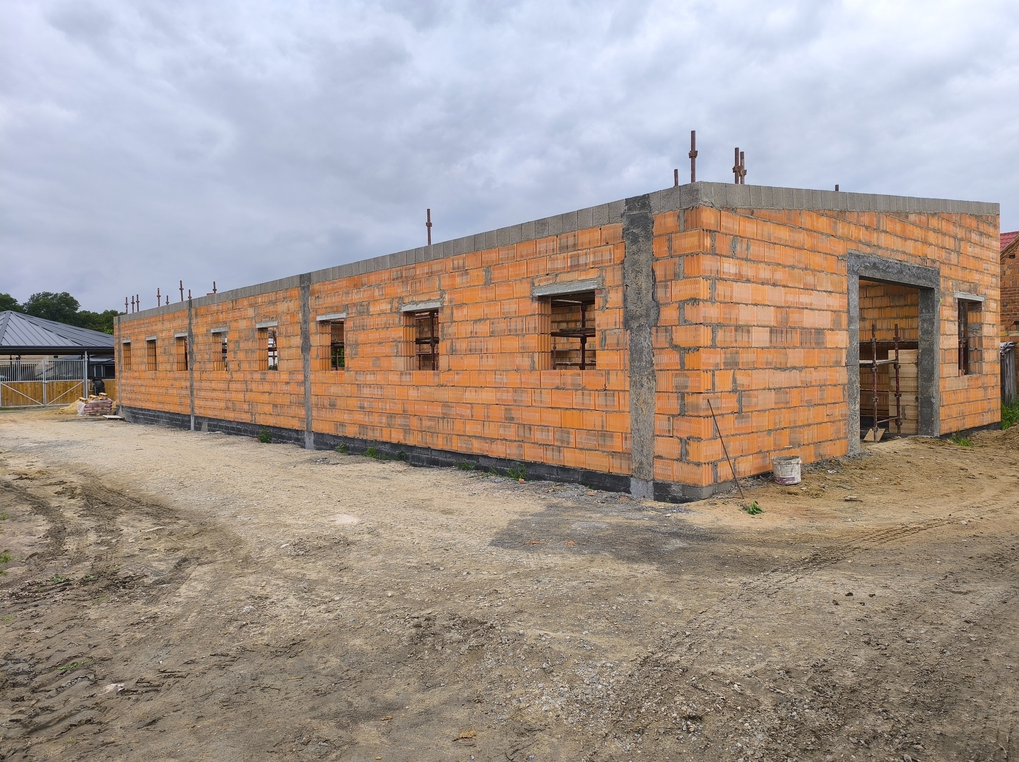 Budowa domu z czerwonej cegły: ściany wzniesione, otwory okienne przygotowane, widoczne zbrojenia. Tło: pochmurne niebo, teren budowy.