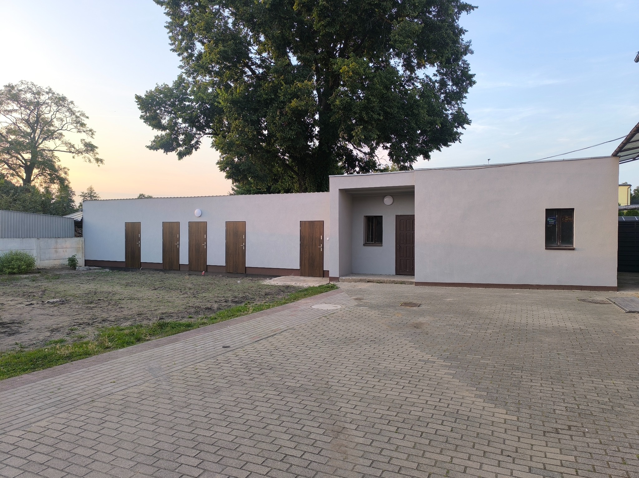 Nowoczesny budynek gospodarczy z jasną elewacją i rzędem brązowych drzwi, otoczony kostką brukową i zielenią, na tle drzew i błękitnego nieba.