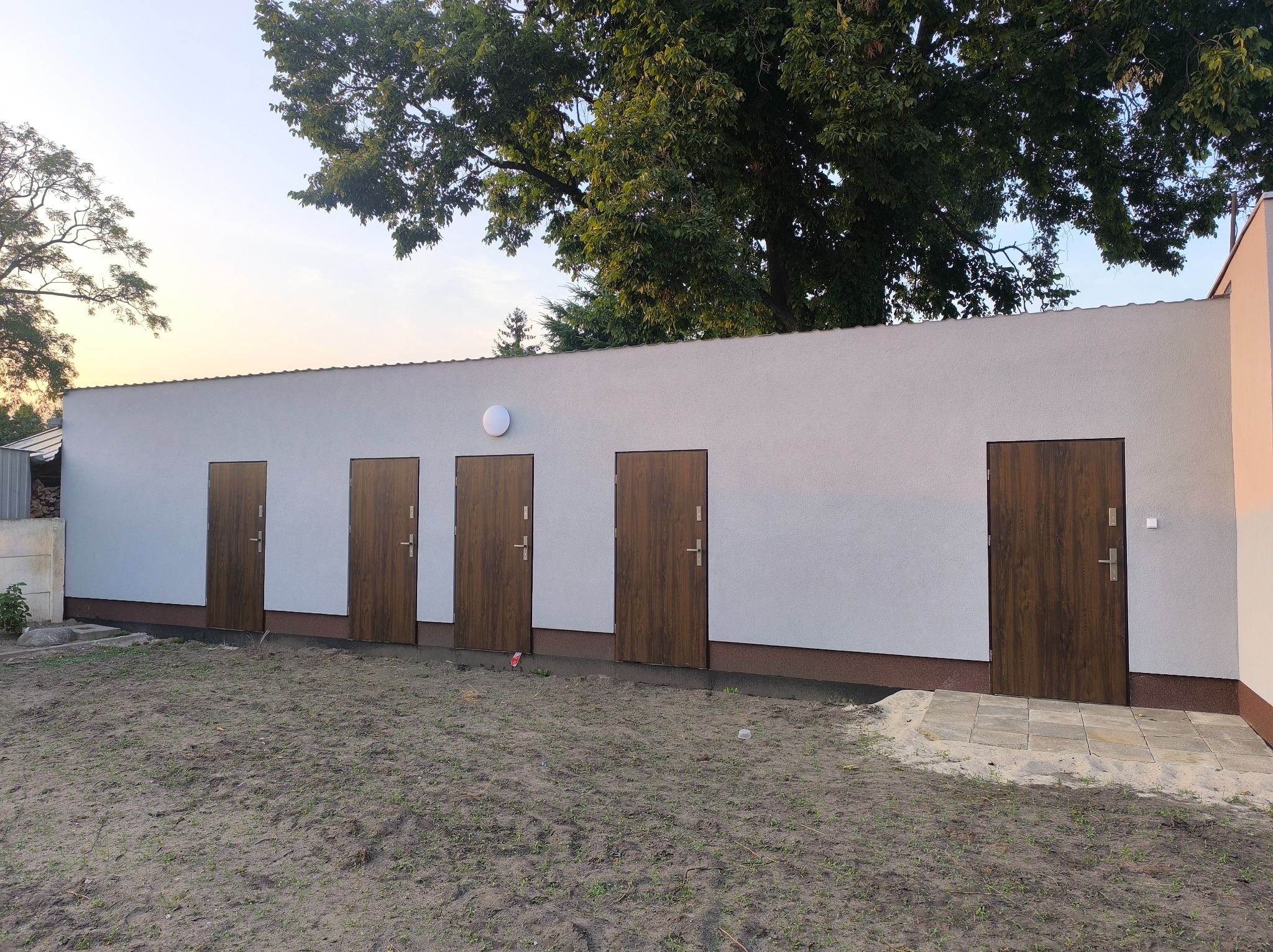 Nowoczesny, parterowy budynek z czterema brązowymi drzwiami wejściowymi i oświetleniem zewnętrznym, otoczony zielenią i drzewami w pogodny dzień.