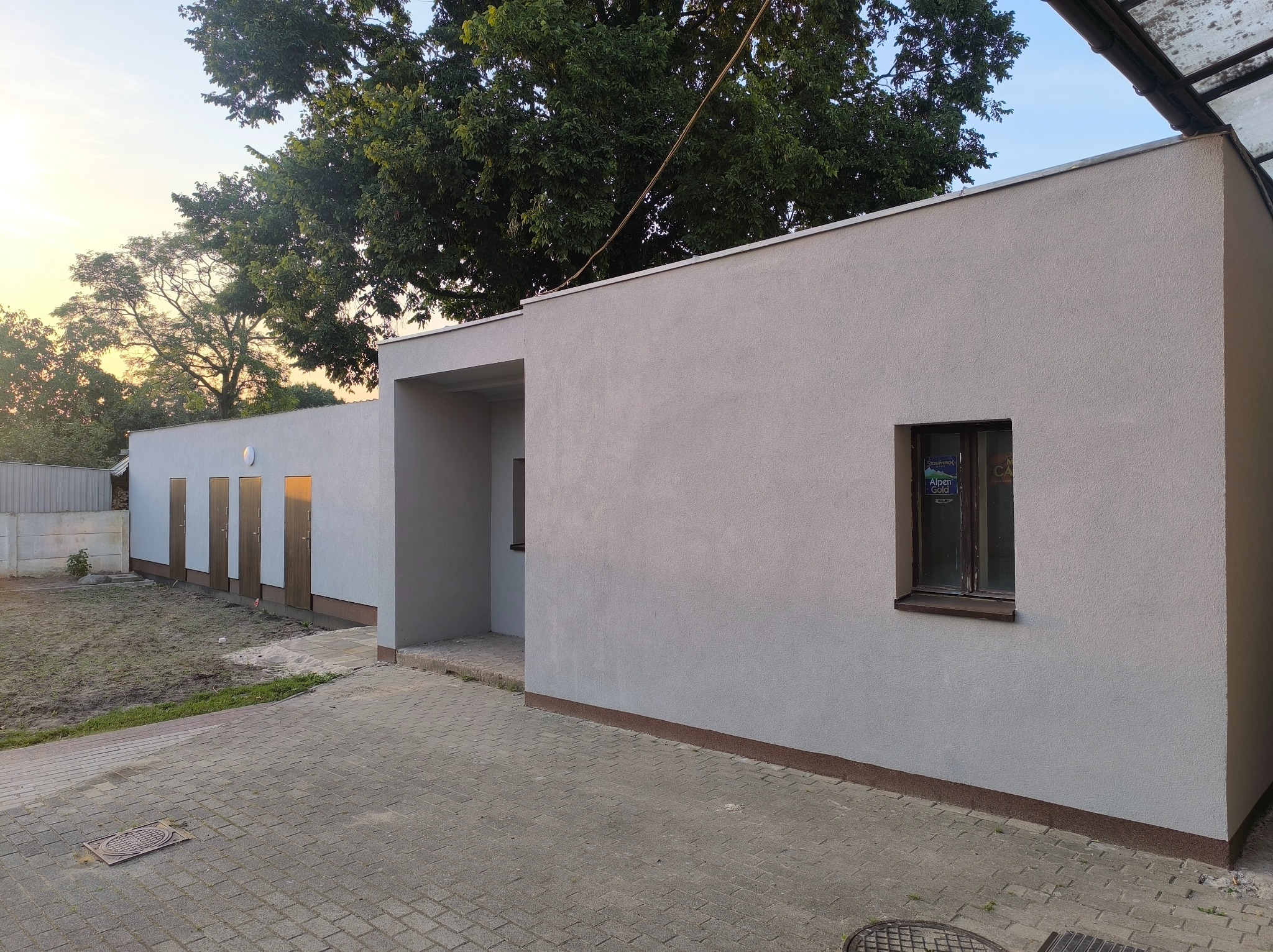 Szara elewacja budynku z brązowymi drzwiami i oknem, widoczna z poziomu chodnika z kostki brukowej. U góry widoczne drzewa i fragment nieba o zachodzie słońca.