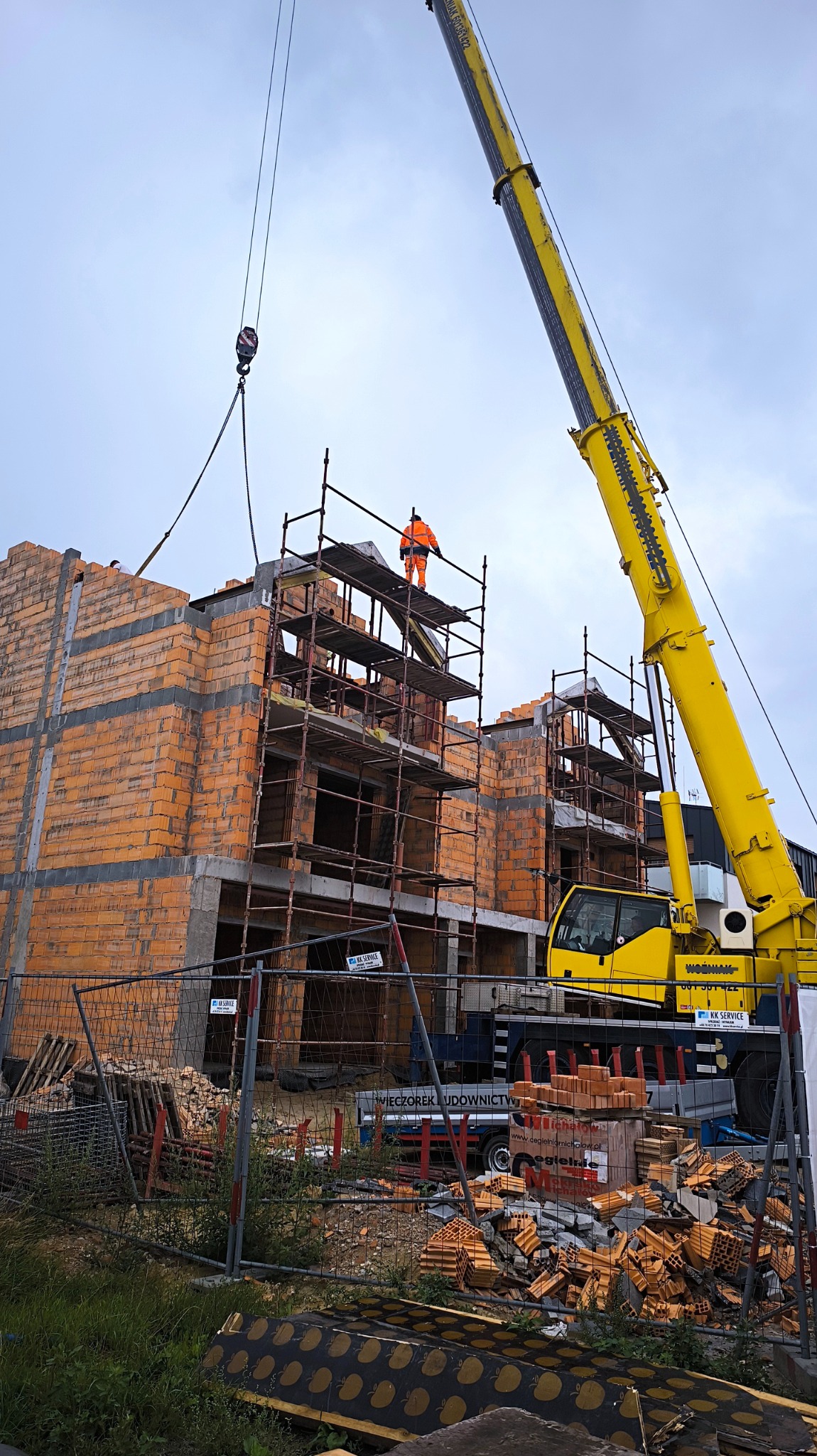 Budowa domu z cegły ceramicznej: murarz w pomarańczowym kombinezonie na rusztowaniu, dźwig budowlany w tle. Widoczne materiały budowlane i ogrodzenie placu budowy.