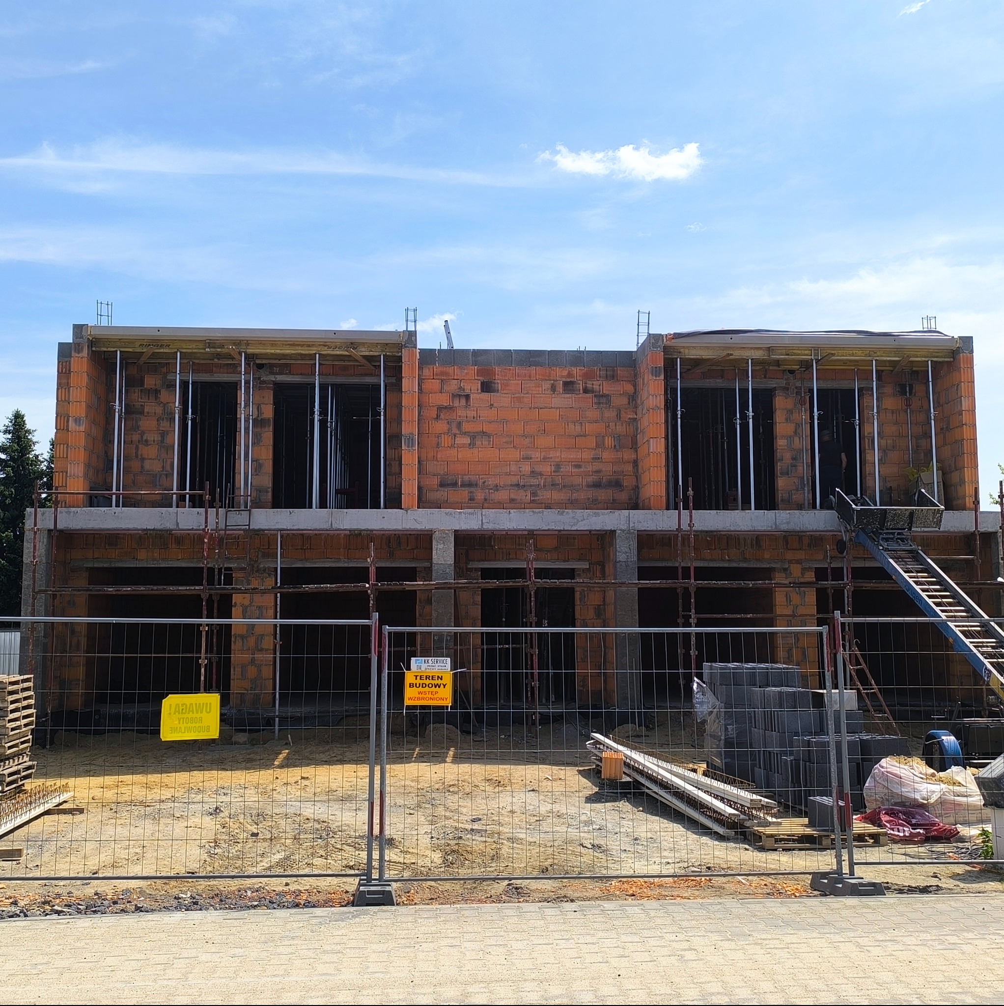Budowa dwukondygnacyjnego domu z czerwonej cegły, widok zza ogrodzenia budowlanego. Widoczne rusztowania i materiały budowlane. Teren budowy zabezpieczony.