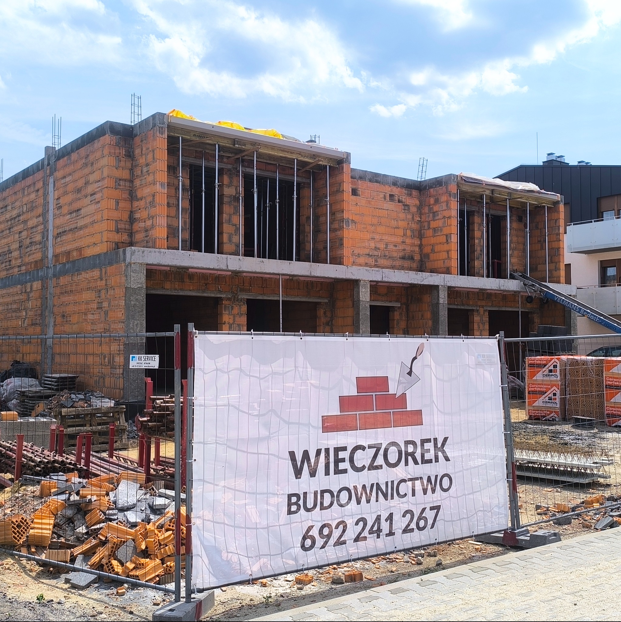 Wznoszenie dwukondygnacyjnego domu z czerwonej cegły. Widoczny baner reklamowy firmy budowlanej 'Wieczorek' i materiały budowlane. Częściowo pochmurne niebo.