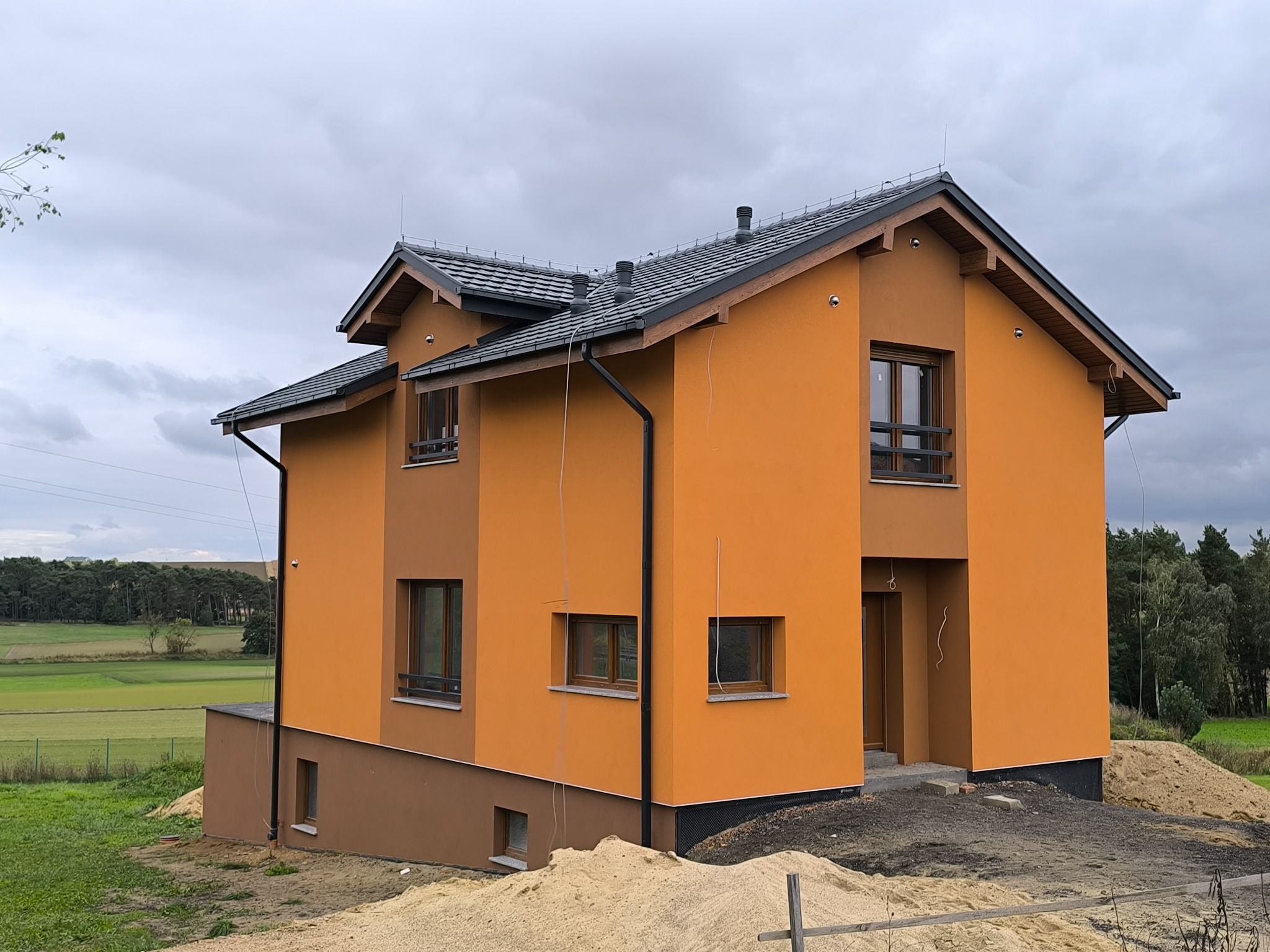 Dwukondygnacyjny dom z pomarańczową elewacją i brązowymi akcentami, ciemnoszarym dachem. Widoczne okna z drewnianymi ramami i otoczenie w trakcie zagospodarowywania.