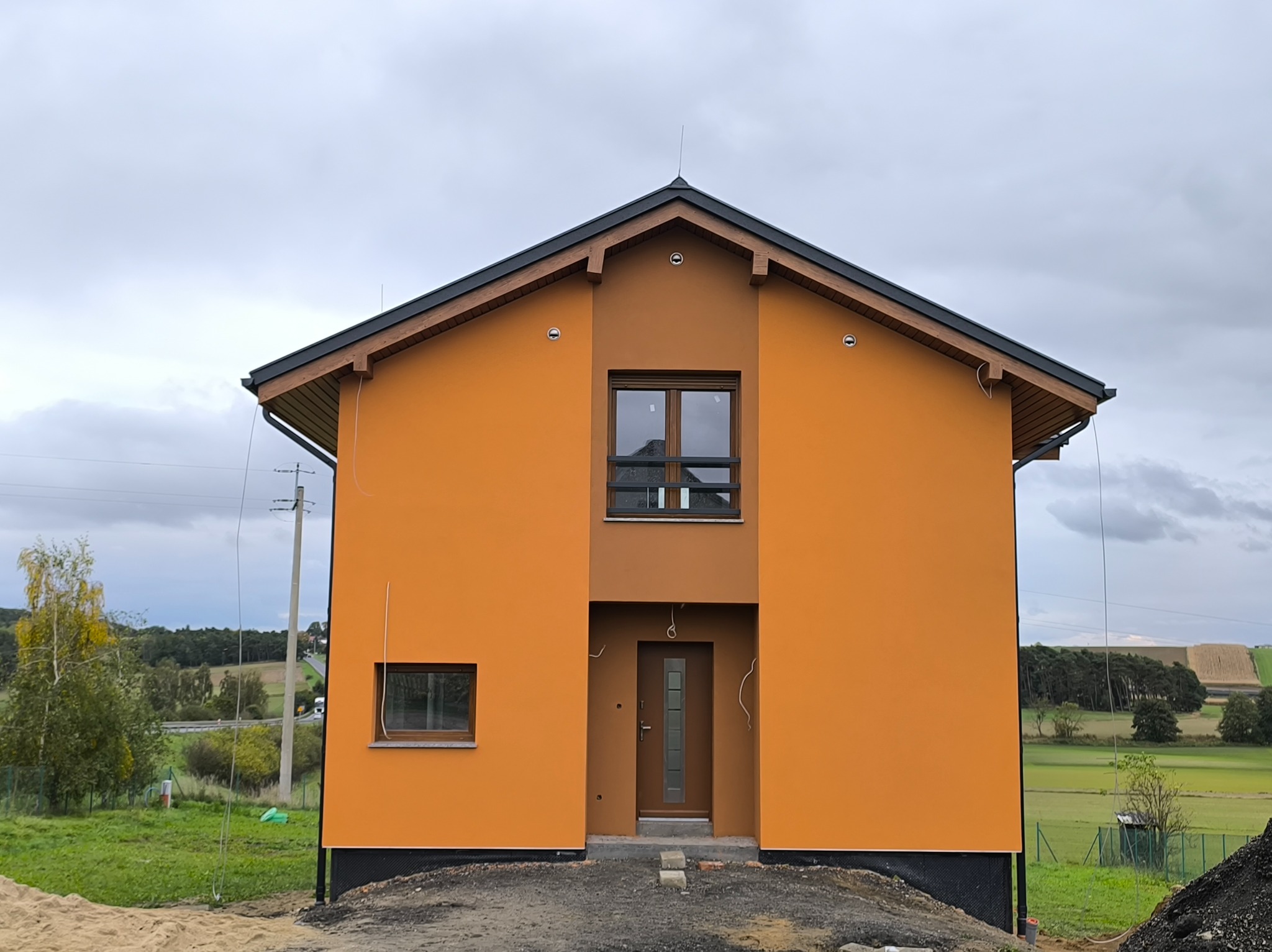 Dwukondygnacyjny dom z pomarańczową elewacją, brązowymi detalami i ciemnym dachem. Widoczne okna, drzwi wejściowe i fragment otoczenia z zielenią. Pochmurne niebo w tle.