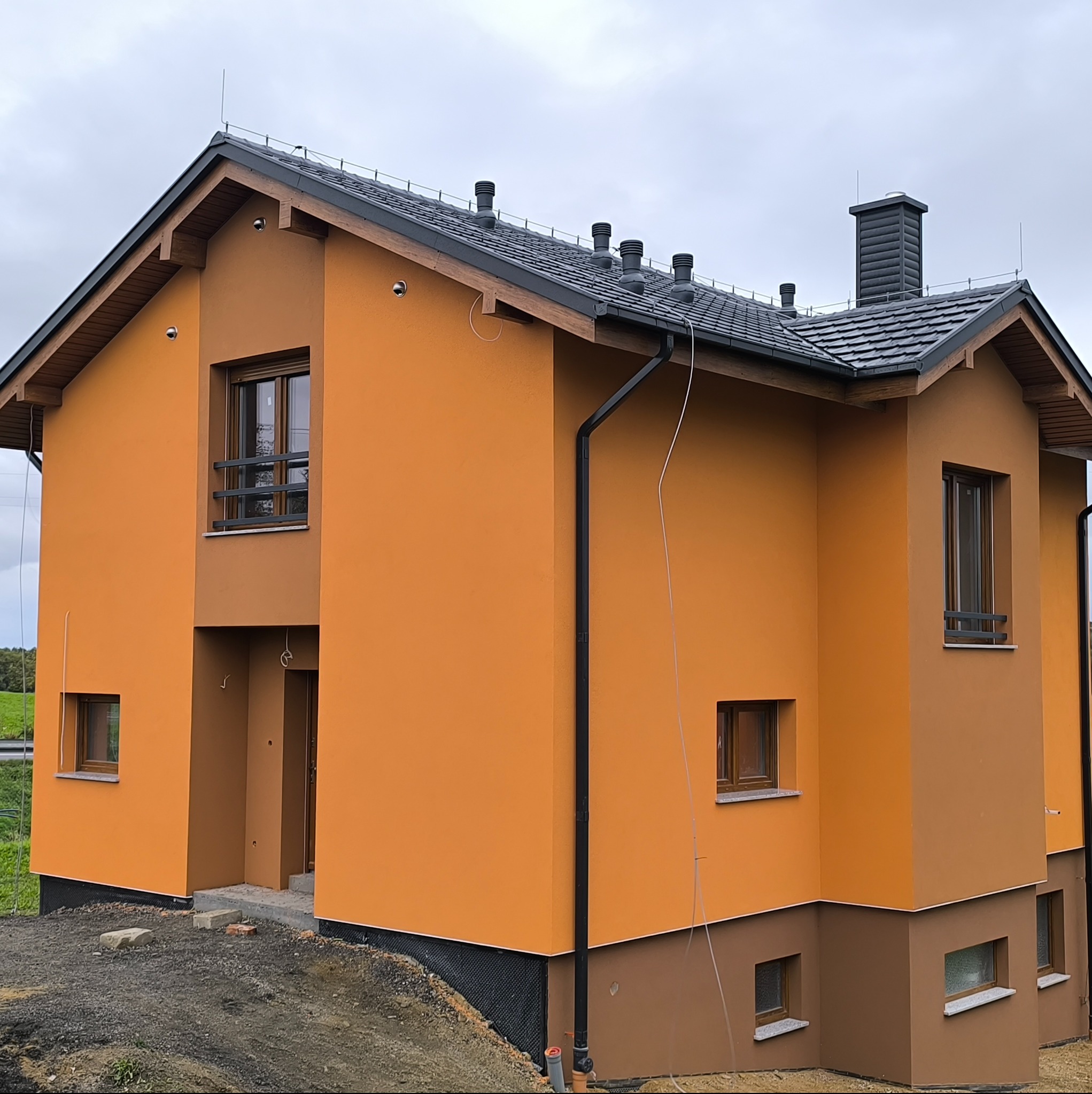 Dwukondygnacyjny dom z pomarańczową elewacją i ciemną dachówką, widoczny z perspektywy narożnej. Na dachu komin i wentylatory. Budynek na nierównym terenie.