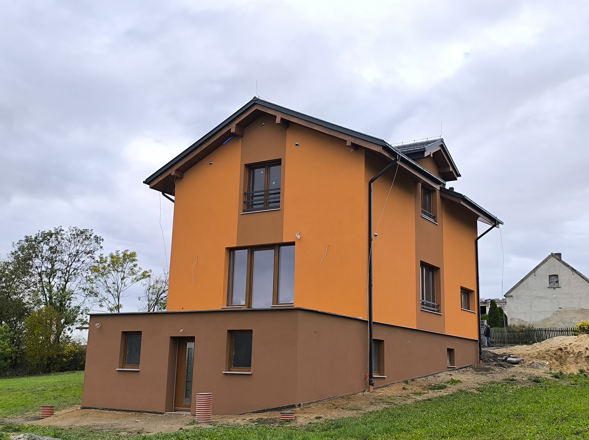 Dwukondygnacyjny dom z pomarańczową elewacją i brązowym cokołem, widoczny z perspektywy trawnika. W tle drzewa i sąsiedni budynek. Pochmurne niebo.