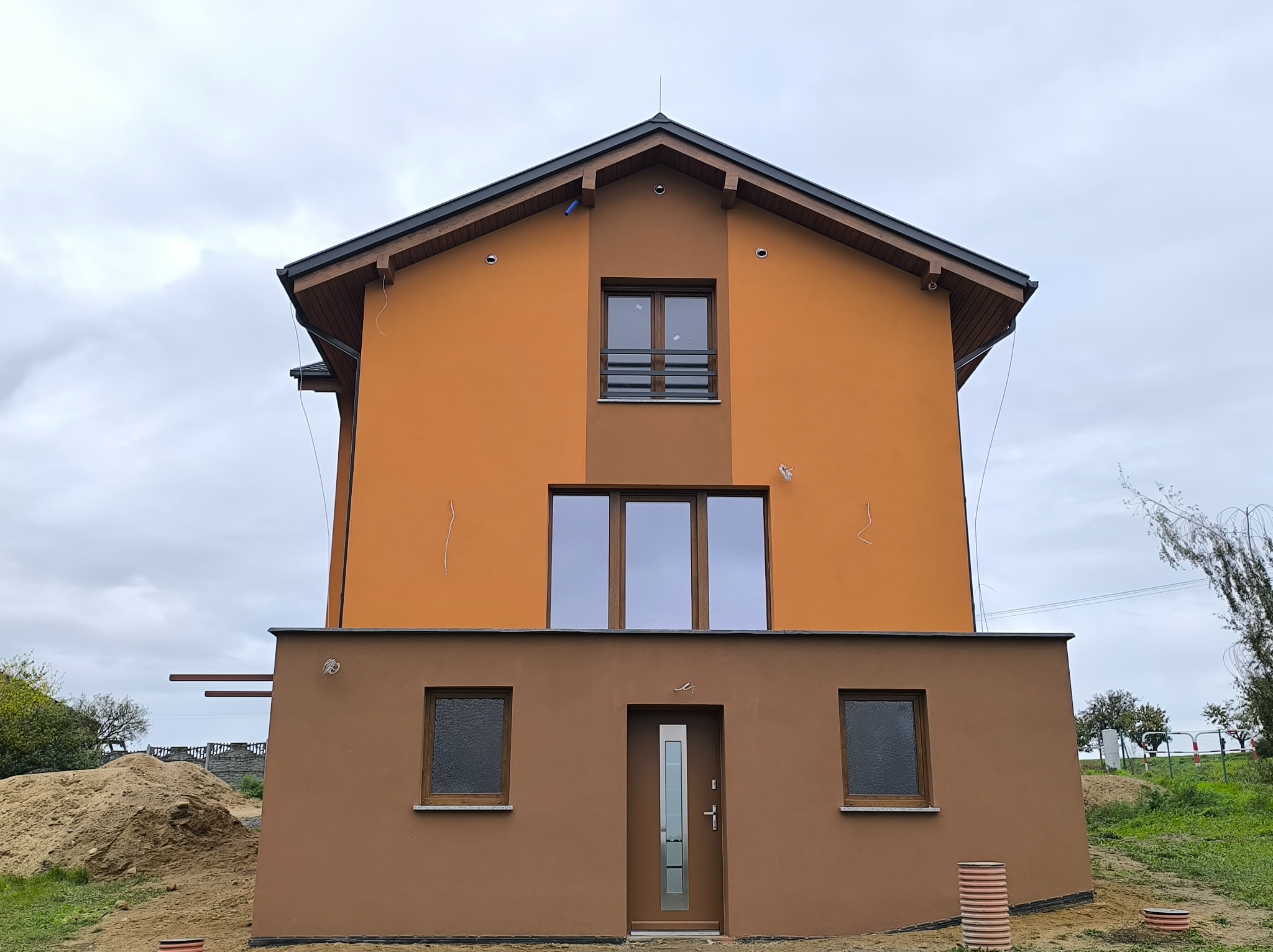 Dwukondygnacyjny dom z elewacją w odcieniach brązu i pomarańczy, z widocznym stosem piasku obok, sugerujący prace budowlane w toku.