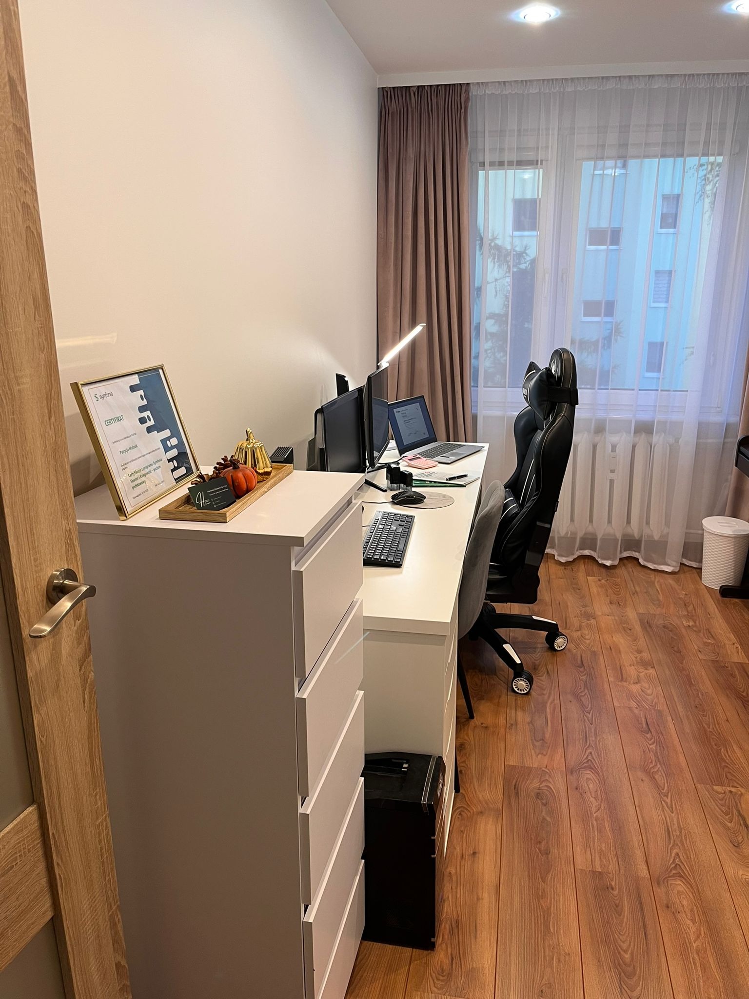 Biuro domowe z białym biurkiem, laptopem, certyfikatem i dekoracjami. W tle okno z firankami i widok na budynek. Ergonomiczne krzesło biurowe.