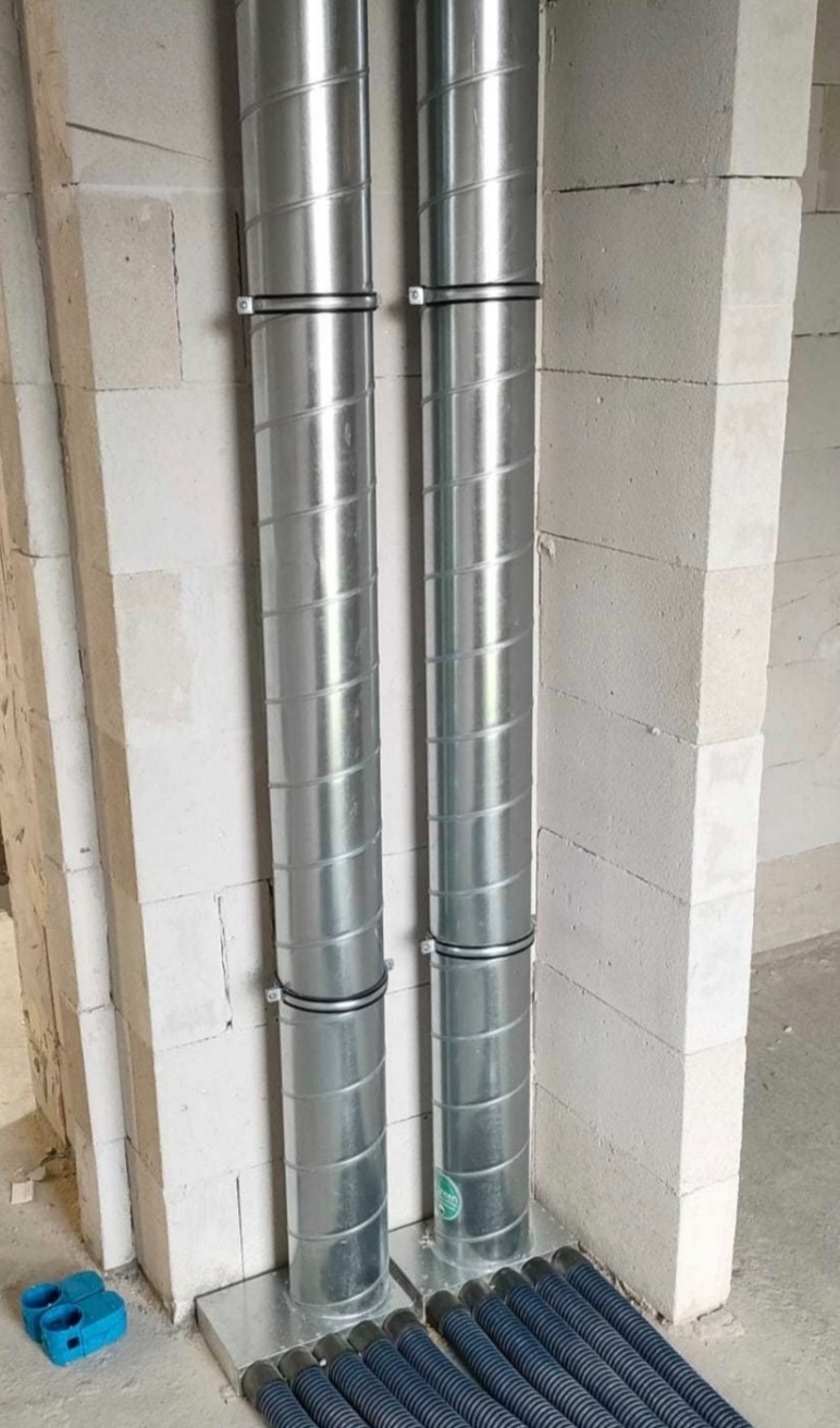 Dwie pionowe, spiralne rury wentylacyjne ze stali nierdzewnej, zamocowane do ściany z białych bloczków, z widocznymi niebieskimi złączkami i niebieskimi rurami karbowanymi na podłodze.