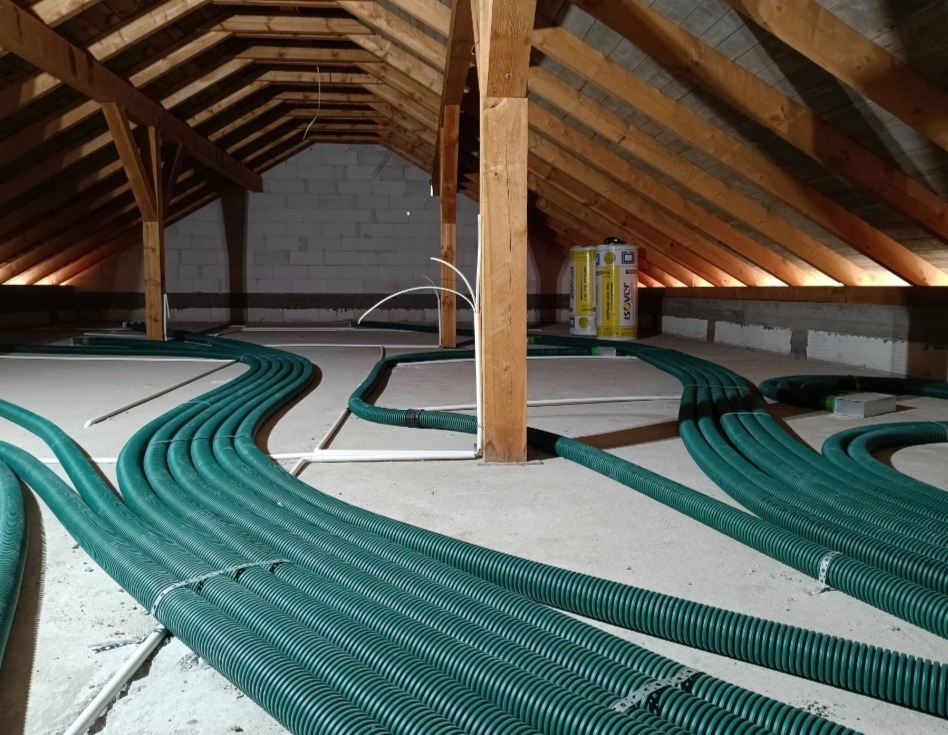 Zielone rury wentylacyjne ułożone na poddaszu z drewnianą konstrukcją dachu i ociepleniem, widoczne puszki izolacyjne Isover