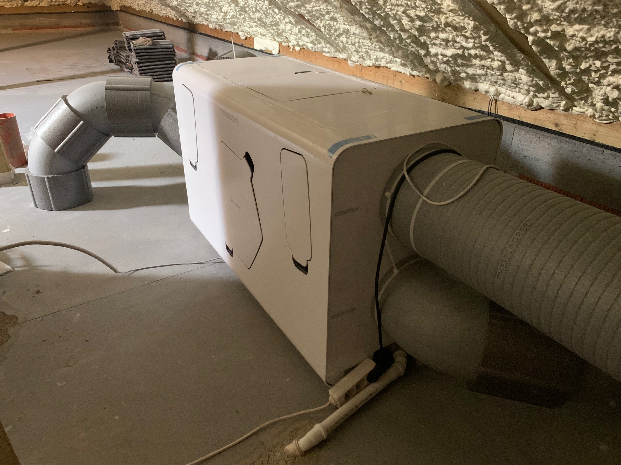 Biała jednostka centralna wentylacyjna z podłączonymi szarymi przewodami wentylacyjnymi w surowym pomieszczeniu na poddaszu z izolacją natryskową na skosach.