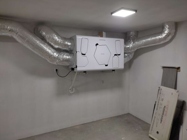 Zamontowany rekuperator z podłączonymi, izolowanymi przewodami wentylacyjnymi w surowym pomieszczeniu z widocznym oświetleniem sufitowym.