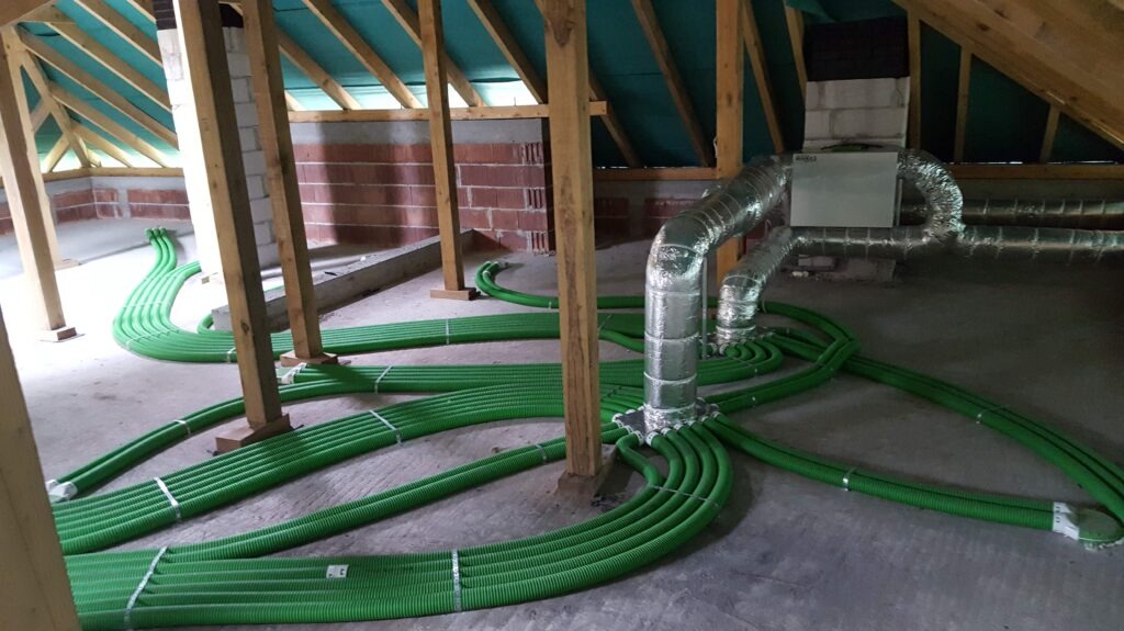 Montaż instalacji wentylacyjnej z zielonymi rurami w surowym wnętrzu poddasza, widoczny centralny rekuperator i aluminiowe przewody wentylacyjne.