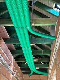 Zielone, karbowane rury wentylacyjne prowadzone pod drewnianą konstrukcją dachu w surowym stanie, widoczne ceglane ściany.