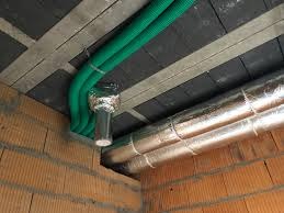 Zielone, karbowane rury wentylacyjne przymocowane do sufitu z szarej płyty, połączone aluminiową taśmą z krótkim, przezroczystym kanałem, widoczne również izolowane rury wentylacyjne i fragment...