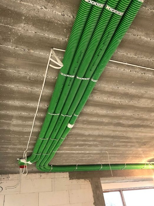 Montaż instalacji wentylacyjnej: wiązka zielonych, karbowanych rur przymocowanych do surowego betonowego stropu za pomocą metalowych uchwytów.
