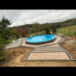 TURBUD - Owalny basen z błękitną wodą otoczony nową, dwukolorową kostką brukową prowadzącą do schodów z kostki, na tle zielonego krajobrazu i pochmurnego nieba.