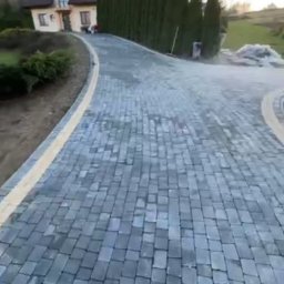 TURBUD - Szeroki kadr na podjazd z szarej kostki brukowej prowadzący do domu z brązowym dachem, obok żywopłot z tui, w tle błękitne niebo z białymi smugami.