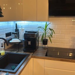 TURBUD - Nowoczesna kuchnia z białymi płytkami typu metro, czarnym zlewem i armaturą, drewnianym blatem, podświetleniem LED pod szafkami oraz sprzętami AGD: ekspres do kawy, robot kuchenny i płyta indukcyjna.