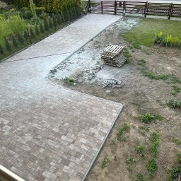 TURBUD - Układanie kostki brukowej w ogrodzie na posesji prywatnej, widok z góry na częściowo ułożoną nawierzchnię z paletami kostki i przygotowanym podłożem.