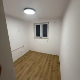 Pusty pokój z panelami podłogowymi w odcieniach brązu, białe ściany z widocznymi rurami i wystającym kablem, okno z białymi ramami i okrągła lampa sufitowa.