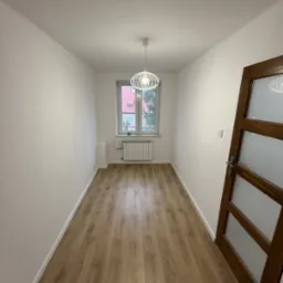 Wąski pokój po remoncie z panelami podłogowymi w odcieniach brązu, białe ściany, okno z grzejnikiem i nowoczesną lampą wiszącą.