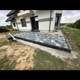 Nowo ułożony taras z szarej kostki brukowej przy nowoczesnym domu z białymi ścianami i szarymi akcentami, widoczny wózek i zagęszczarka w tle.