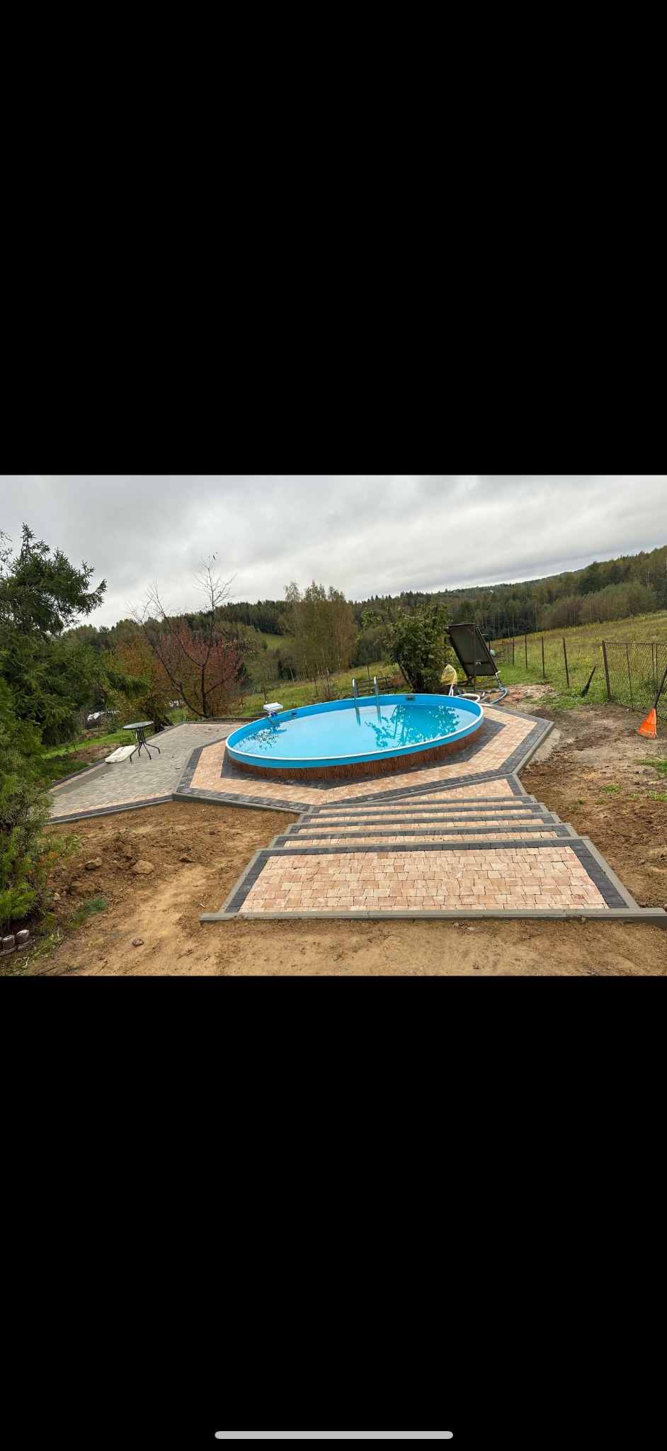 Owalny basen z błękitną wodą otoczony nową, dwukolorową kostką brukową prowadzącą do schodów z kostki, na tle zielonego krajobrazu i pochmurnego nieba.