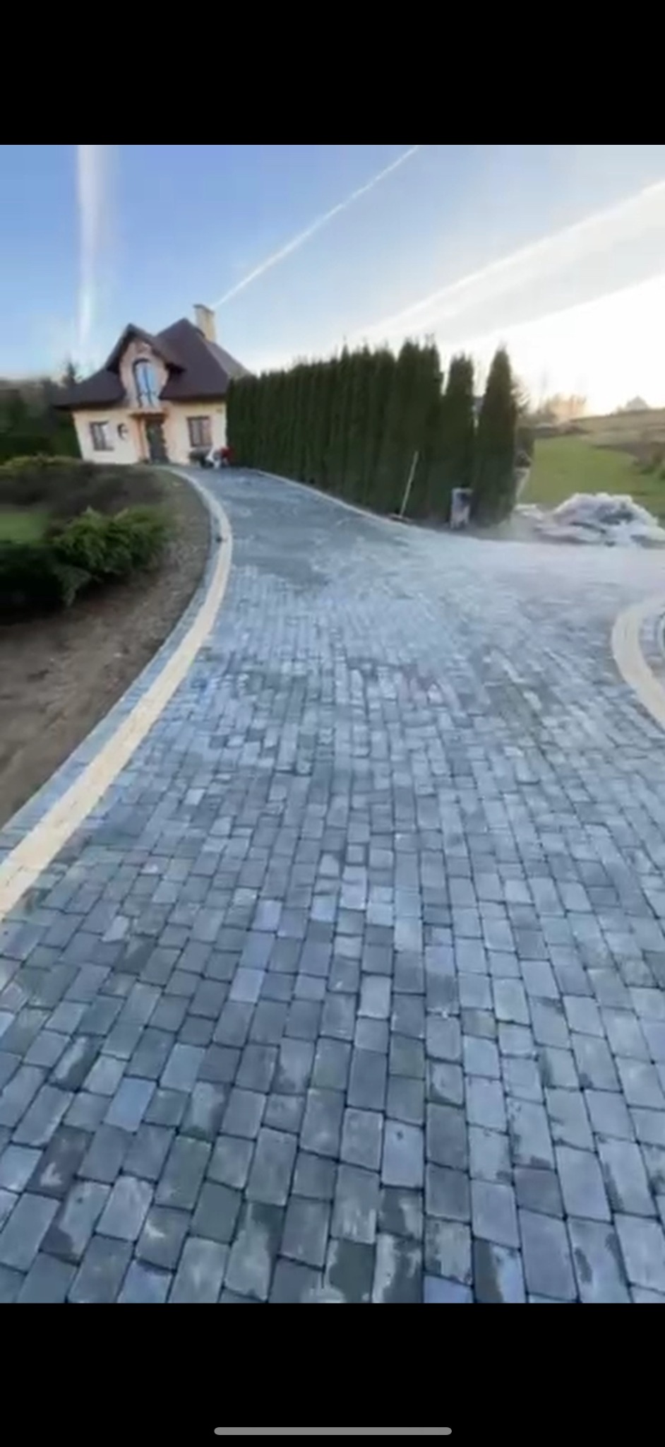 Szeroki kadr na podjazd z szarej kostki brukowej prowadzący do domu z brązowym dachem, obok żywopłot z tui, w tle błękitne niebo z białymi smugami.