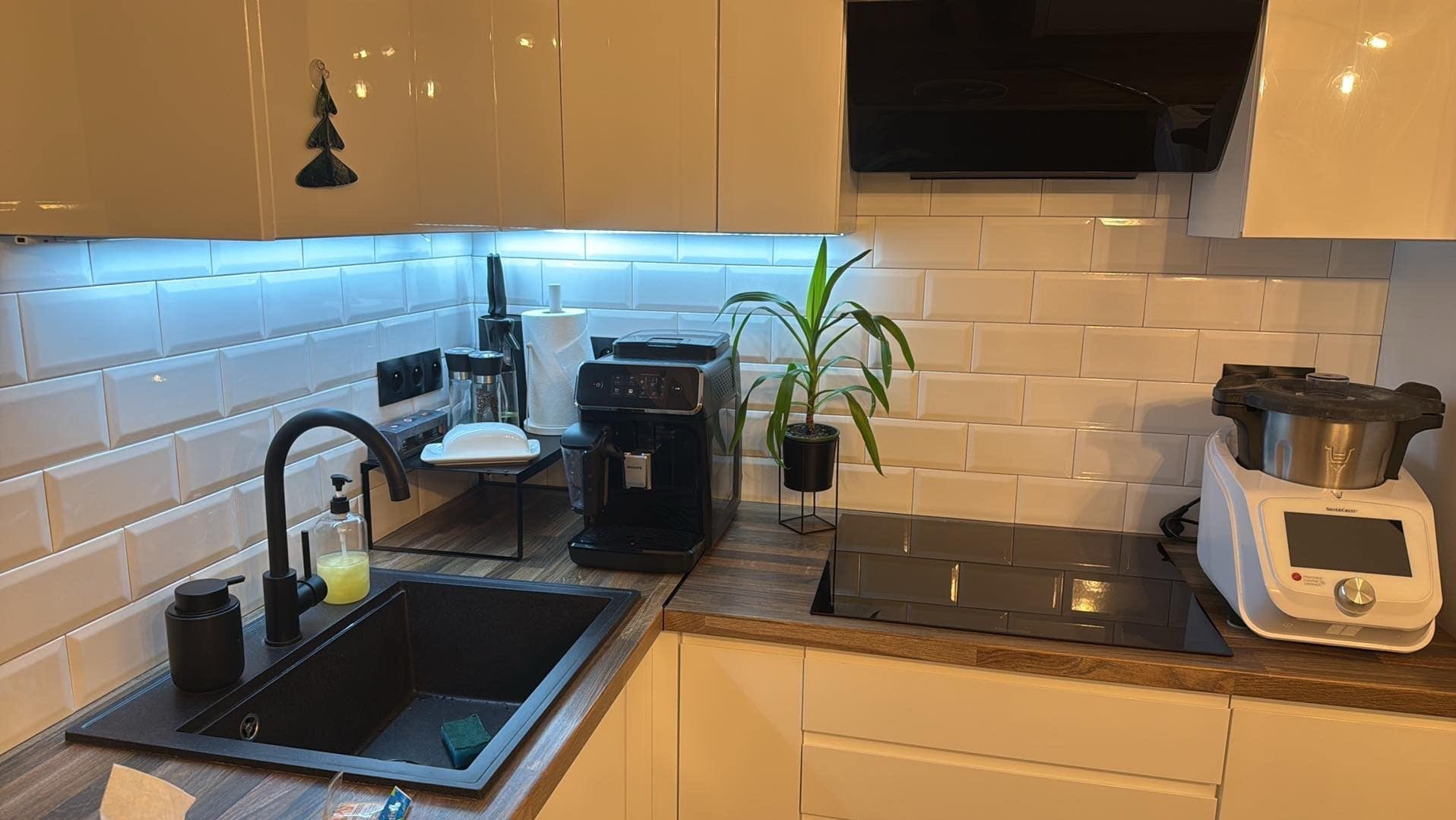 Nowoczesna kuchnia z białymi płytkami typu metro, czarnym zlewem i armaturą, drewnianym blatem, podświetleniem LED pod szafkami oraz sprzętami AGD: ekspres do kawy, robot kuchenny i płyta indukcyjna.