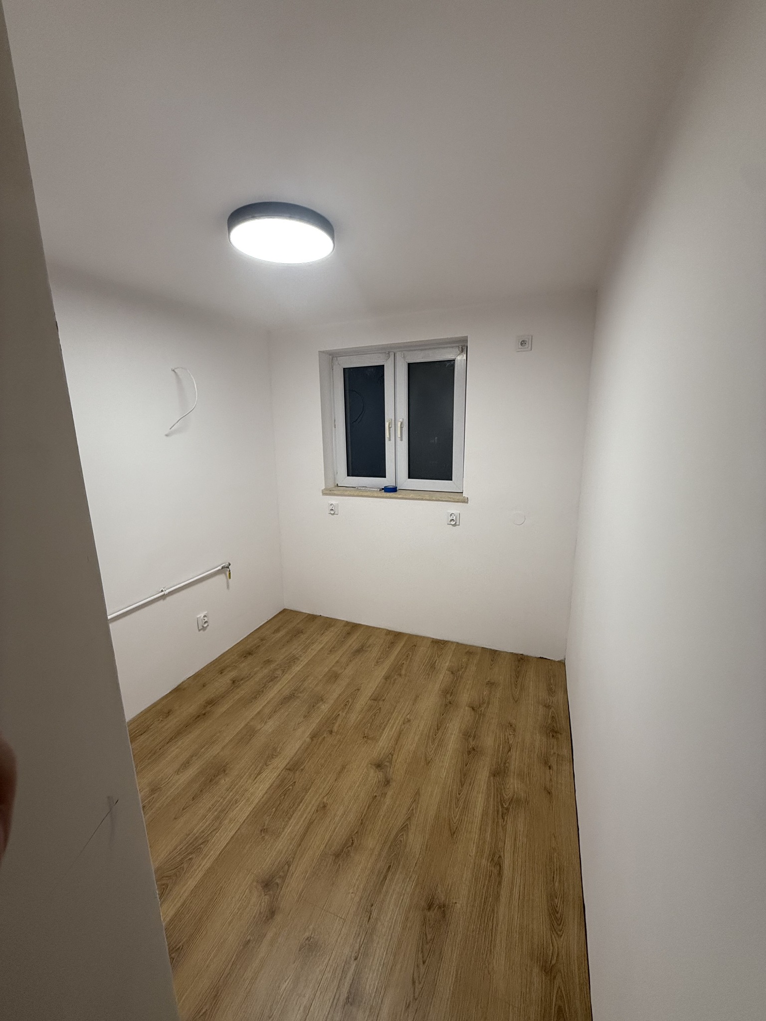 Pusty pokój z panelami podłogowymi w odcieniach brązu, białe ściany z widocznymi rurami i wystającym kablem, okno z białymi ramami i okrągła lampa sufitowa.