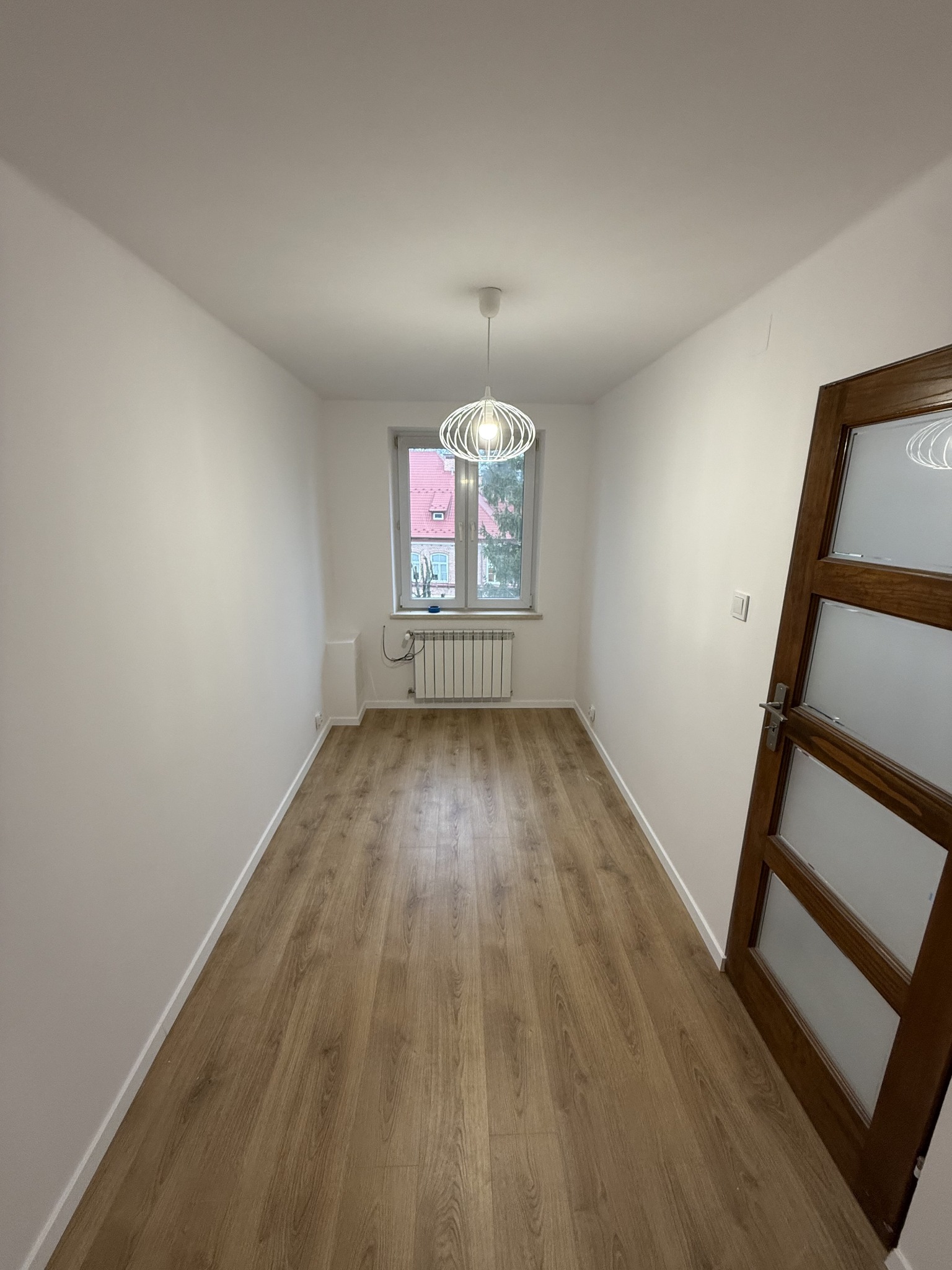 Wąski pokój po remoncie z panelami podłogowymi w odcieniach brązu, białe ściany, okno z grzejnikiem i nowoczesną lampą wiszącą.