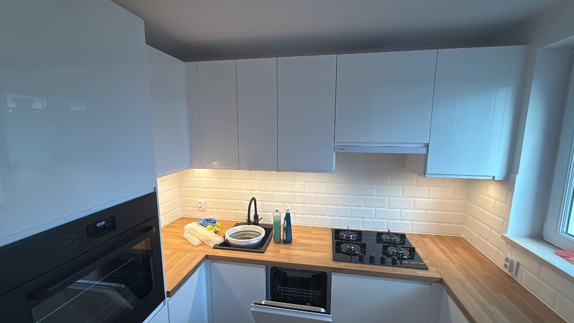 Nowoczesna kuchnia z białymi, lakierowanymi szafkami, drewnianym blatem, czarną płytą grzewczą i białymi płytkami metra pod szafkami; otwarta zmywarka i akcesoria do sprzątania na blacie.