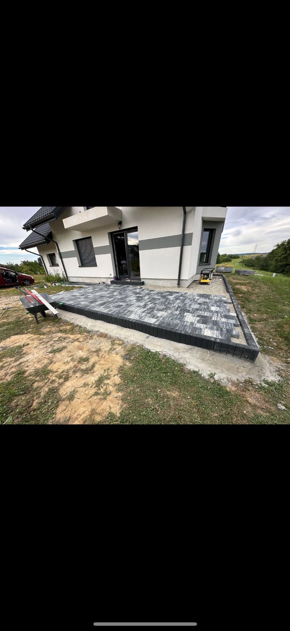 Nowo ułożony taras z szarej kostki brukowej przy nowoczesnym domu z białymi ścianami i szarymi akcentami, widoczny wózek i zagęszczarka w tle.