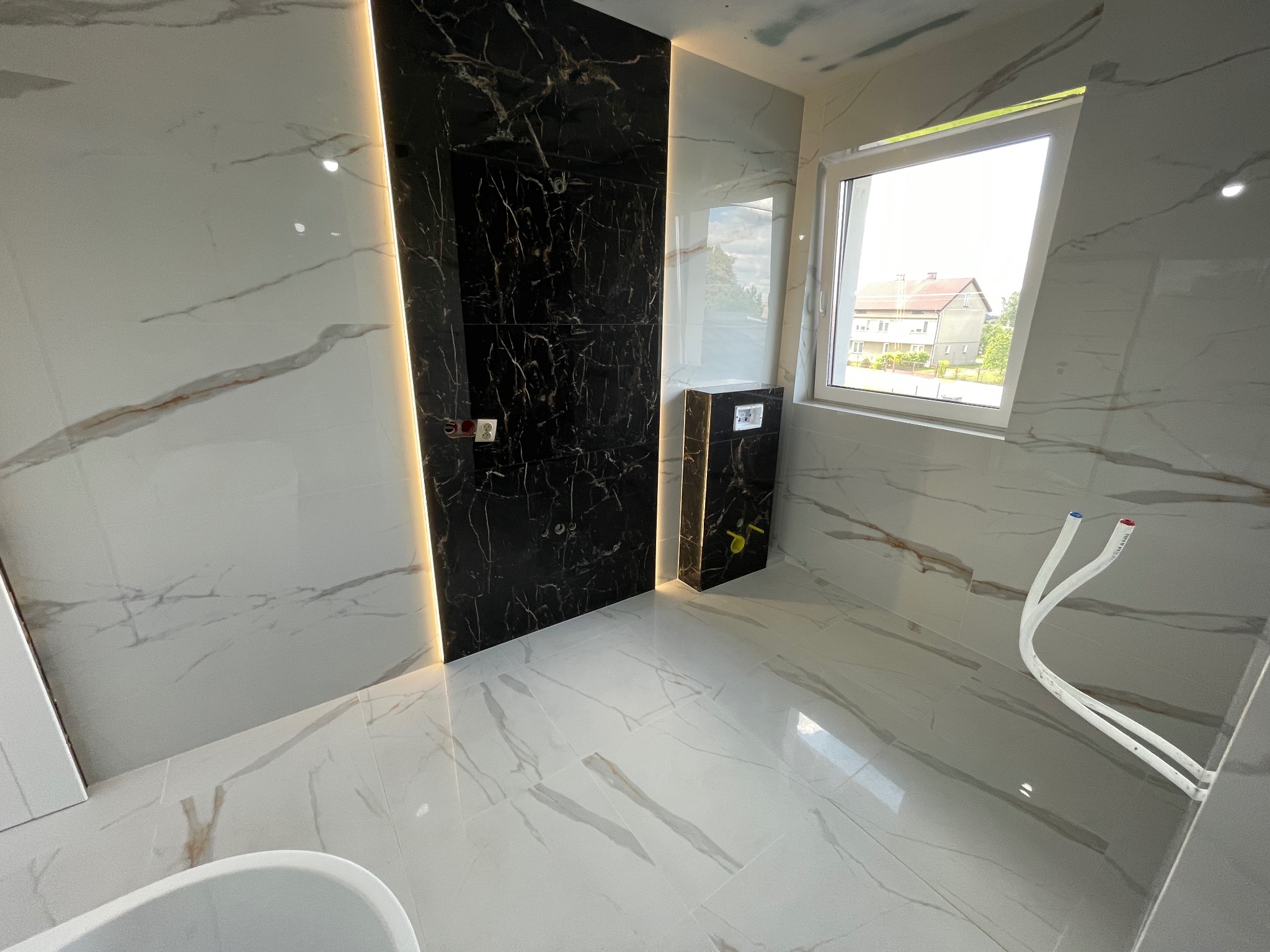 Elegancka łazienka z marmurowymi płytkami, czarnym akcentem i oświetleniem LED. Widoczne przyłącza do armatury i okno z widokiem na budynek.