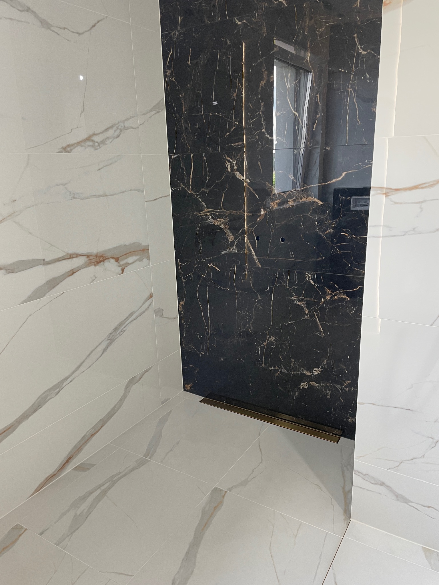 Wykończona ściana i podłoga w łazience z czarnych i białych płytek imitujących marmur. Widoczny odpływ liniowy.