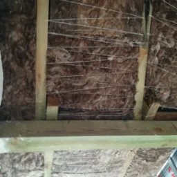 Ocieplenie poddasza wełną mineralną, widoczne drewniane belki konstrukcyjne i drut mocujący izolację, fragment konstrukcji metalowej sufitu podwieszanego.
