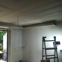 GROTBUDMS SPÓŁKA Z OGRANICZONĄ ODPOWIEDZIALNOŚCIĄ - Wnętrze w trakcie remontu: ściany przygotowane pod malowanie, częściowo zamontowana płyta karton gips na suficie, drabina ustawiona przy ścianie z widocznymi śladami starej farby, lampa budowlana...