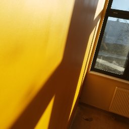 GROTBUDMS SPÓŁKA Z OGRANICZONĄ ODPOWIEDZIALNOŚCIĄ - Ściana pomalowana na intensywny żółty kolor wpadająca w kadr pod kątem, z widocznym oknem w czarnej ramie i grzejnikiem. Silne światło słoneczne tworzy geometryczne cienie na ścianie.