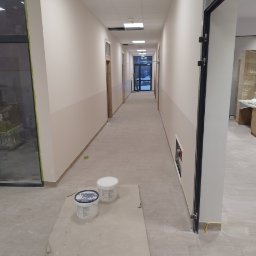 GROTBUDMS SPÓŁKA Z OGRANICZONĄ ODPOWIEDZIALNOŚCIĄ - Długi korytarz w budynku po świeżym pomalowaniu ścian na dwukolorowo, widoczne dwa wiadra z farbą na kartonie zabezpieczającym podłogę.