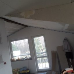 GROTBUDMS SPÓŁKA Z OGRANICZONĄ ODPOWIEDZIALNOŚCIĄ - Poddasze w trakcie remontu: zamontowana płyta karton gips na skosach, widoczne szpachlowanie łączeń i fragment ocieplenia, okna z widokiem na drzewa, elementy garderoby w rogu pomieszczenia.