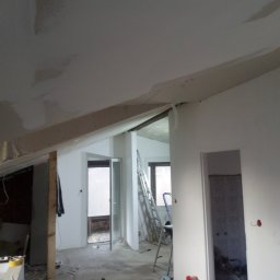 GROTBUDMS SPÓŁKA Z OGRANICZONĄ ODPOWIEDZIALNOŚCIĄ - Poddasze w trakcie prac wykończeniowych: widoczne ocieplenie, płyty kartonowo-gipsowe na skosach sufitu oraz ściany po wstępnym szpachlowaniu, aluminiowa drabina w tle.