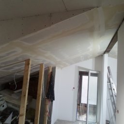 GROTBUDMS SPÓŁKA Z OGRANICZONĄ ODPOWIEDZIALNOŚCIĄ - Skośny sufit poddasza w Łodzi w trakcie montażu płyt karton gips, widoczne drewniane elementy konstrukcyjne i przygotowane płyty. Częściowo nałożona masa szpachlowa.