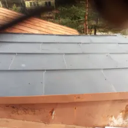 Szare, metalowe panele dachowe, częściowo pomalowane, z widocznymi śrubami mocującymi, sąsiadujące z miedzianym dachem i elementami orynnowania w kolorze rdzy.