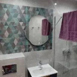 Wykończona łazienka z geometrycznymi płytkami w odcieniach szarości, zieleni i błękitu, okrągłe lustro z oświetleniem LED, biała umywalka na czarnej szafce, szklana kabina prysznicowa z widocznym...