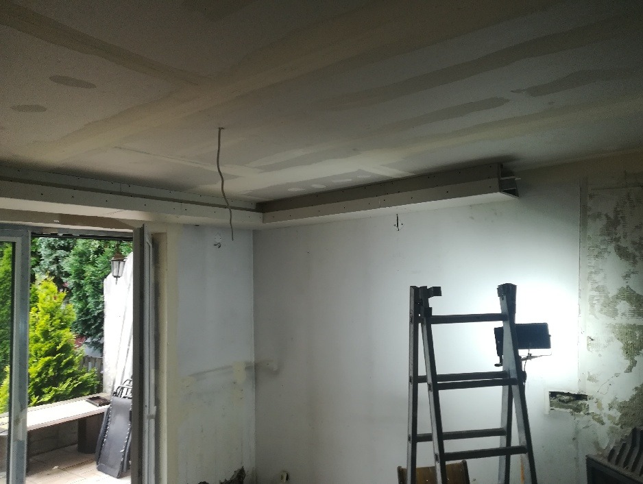 Wnętrze w trakcie remontu: ściany przygotowane pod malowanie, częściowo zamontowana płyta karton gips na suficie, drabina ustawiona przy ścianie z widocznymi śladami starej farby, lampa budowlana...