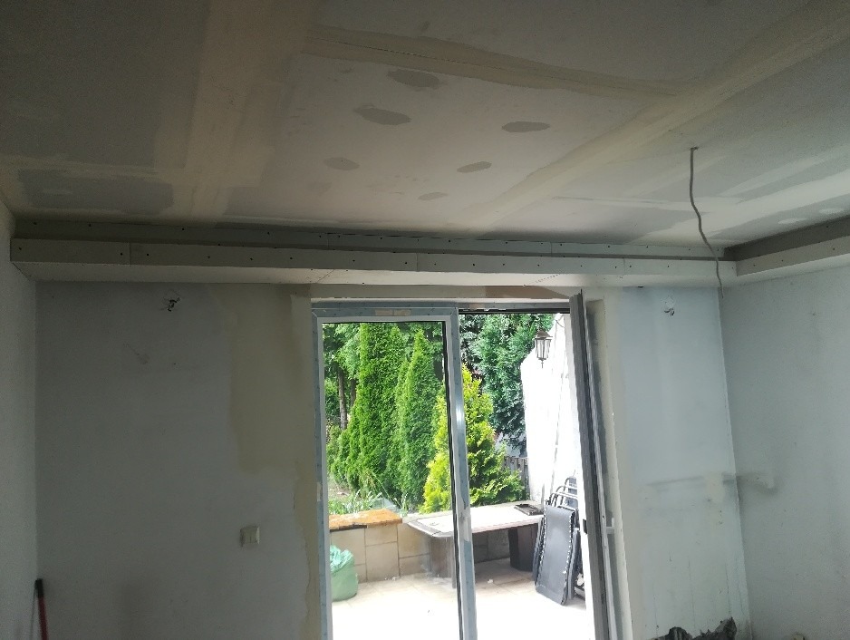 Wnętrze w trakcie prac wykończeniowych: sufit z płyt kartonowo-gipsowych, widoczne łączenia płyt wymagające szpachlowania, ściany częściowo przygotowane pod malowanie, z widokiem na ogród przez...