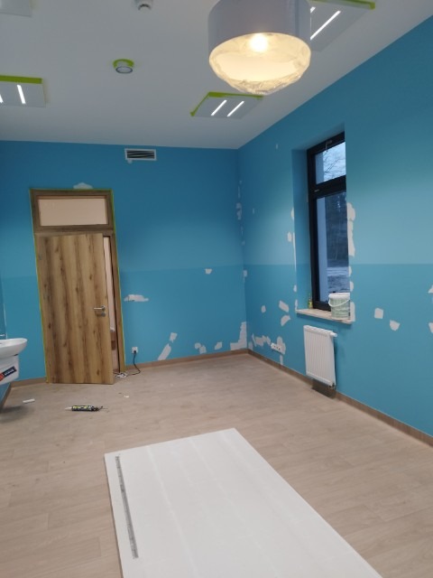 Pokój w trakcie prac wykończeniowych: niebieskie ściany z widocznymi plamami szpachli, drewniane drzwi, biały panel podłogowy i grzejnik pod oknem, lampa sufitowa w folii ochronnej.