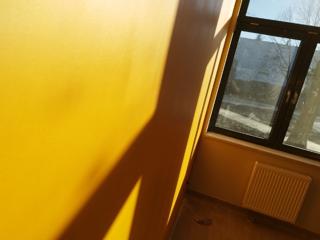 Ściana pomalowana na intensywny żółty kolor wpadająca w kadr pod kątem, z widocznym oknem w czarnej ramie i grzejnikiem. Silne światło słoneczne tworzy geometryczne cienie na ścianie.